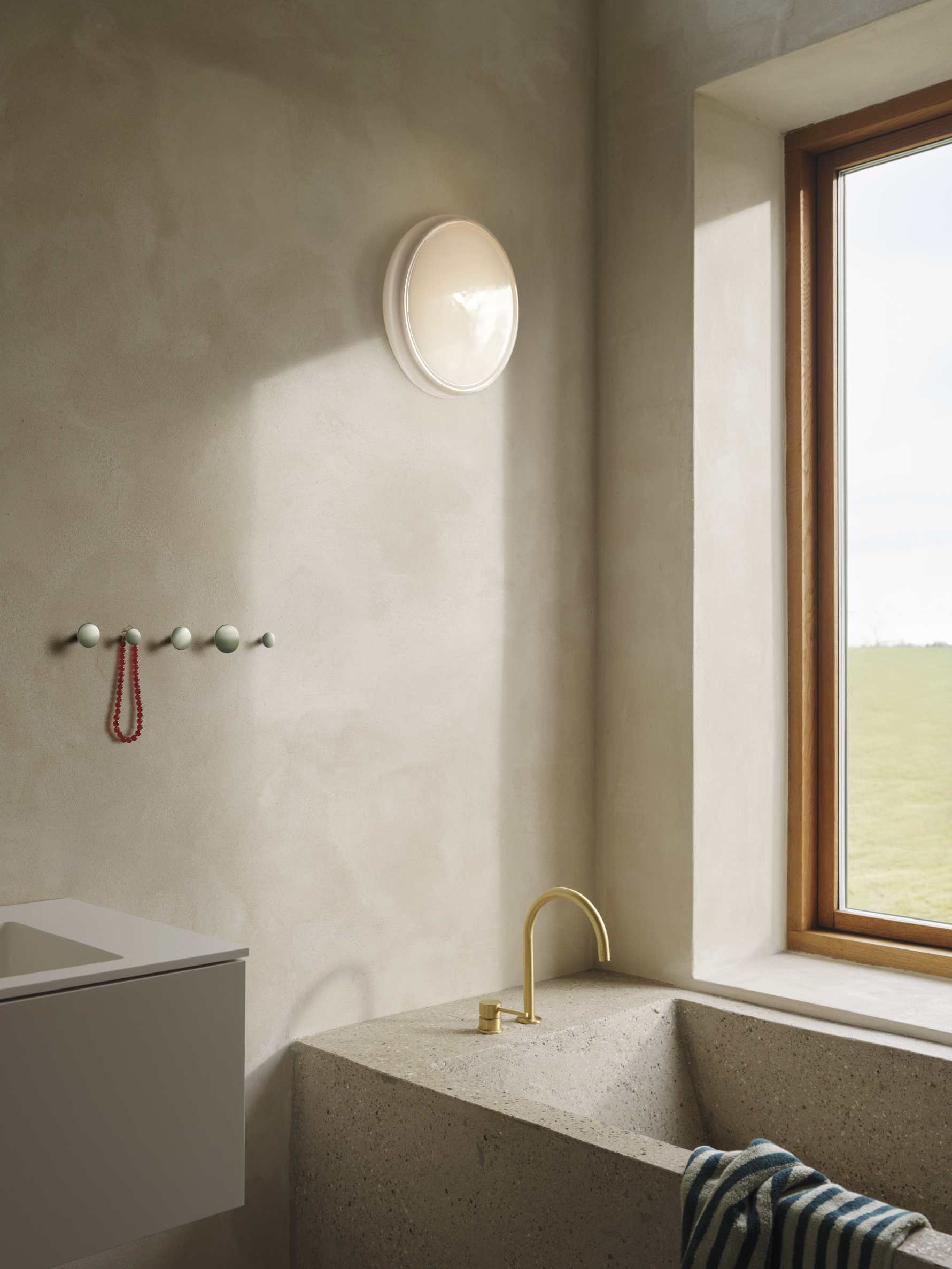 Beige Badezimmer-Szene mit Muuto Looped Wandleuchte, Waschtisch, Armatur und Fenster mit Blick ins Grüne.