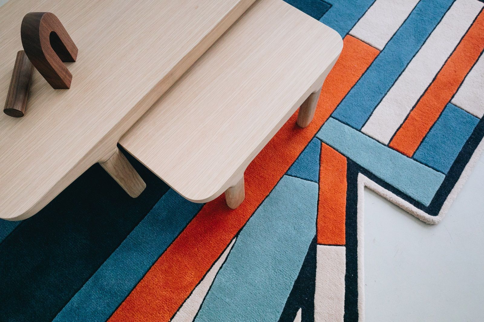Inès Carpet Teppich Hart mit geometrischem Muster in Blau, Orange und Weiß unter einem Holztisch.