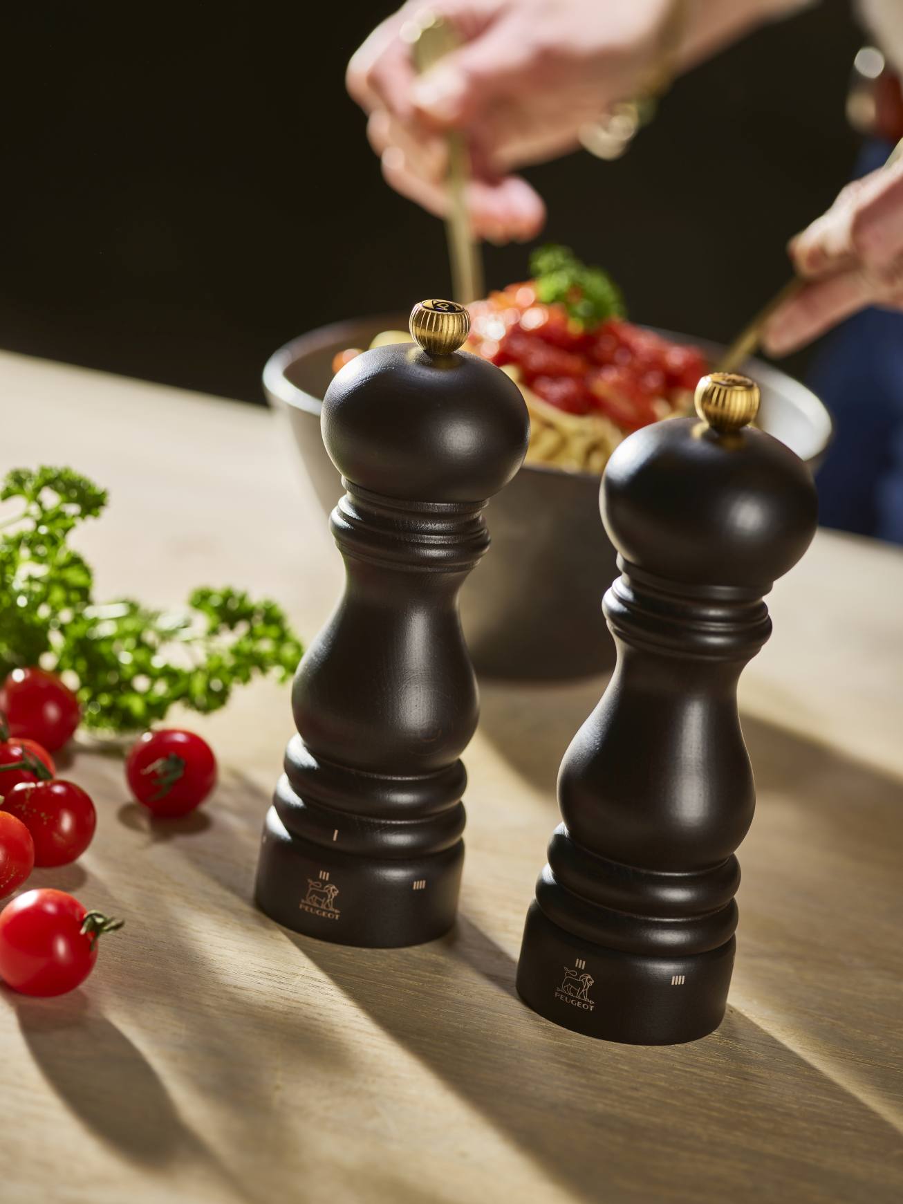 Zwei Peugeot Paris Pfeffermühlen in Schokoladenbraun auf einem Holztisch mit Tomaten und Petersilie.