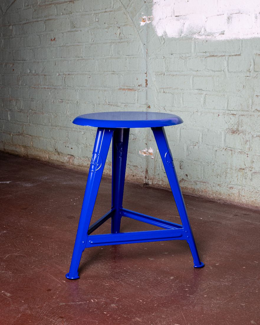 Blauer Rowac Schemel IS 48 cm, ein robuster Industriehocker mit Stahlgestell vor einer Ziegelwand.