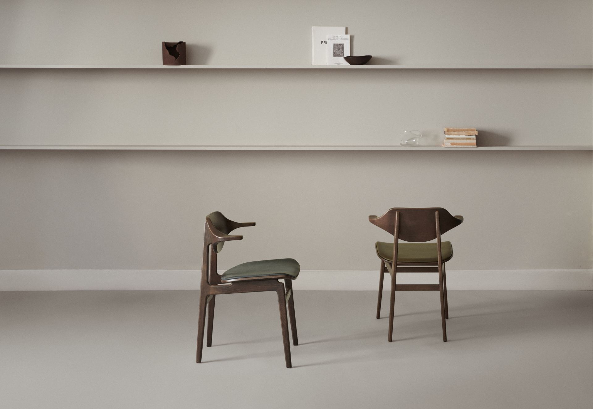 Zwei NORR11 Bufala Dining Stühle aus dunklem Holz mit grünen Sitzen vor Regalen.