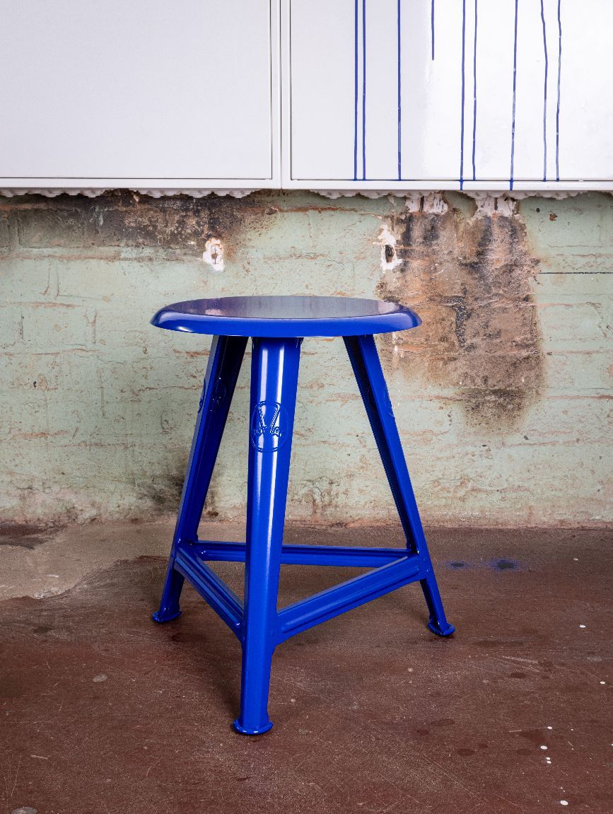 Blauer Rowac Schemel IS, 48 cm hoch, mit Stahlrohrrahmen und runder Sitzfläche vor Wand.