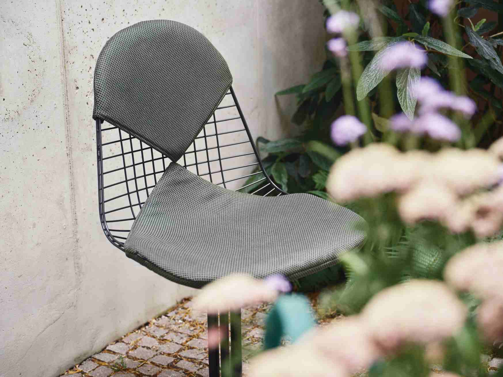 Vitra Wire Chair Bikini Outdoor mit grauem Sitzkissen und schwarzem Drahtgestell vor Pflanzen.