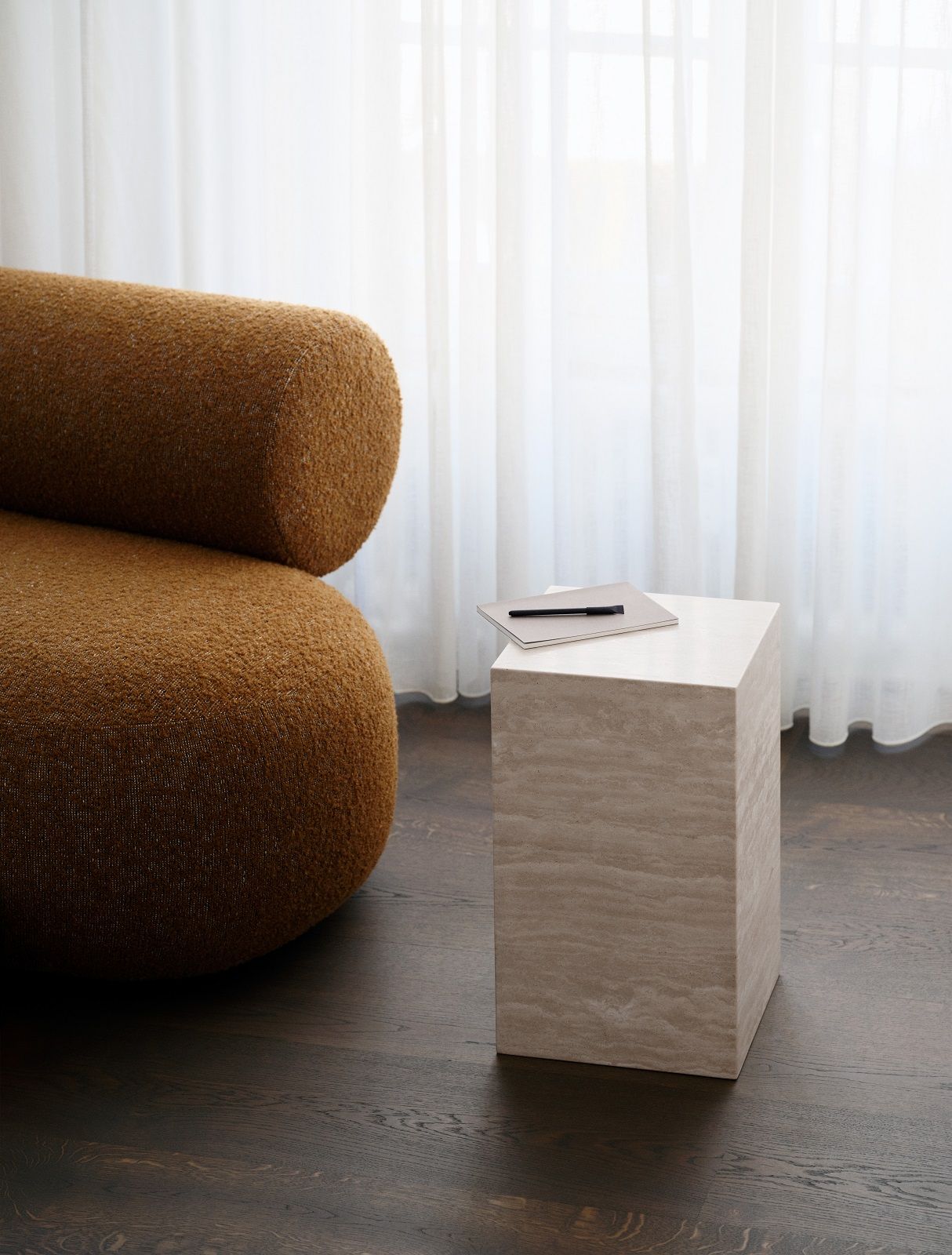 Nahaufnahme des Hippo Sofa von Norr11 in Braun mit Holzhocker, Notizbuch und Stift vor Vorhang.