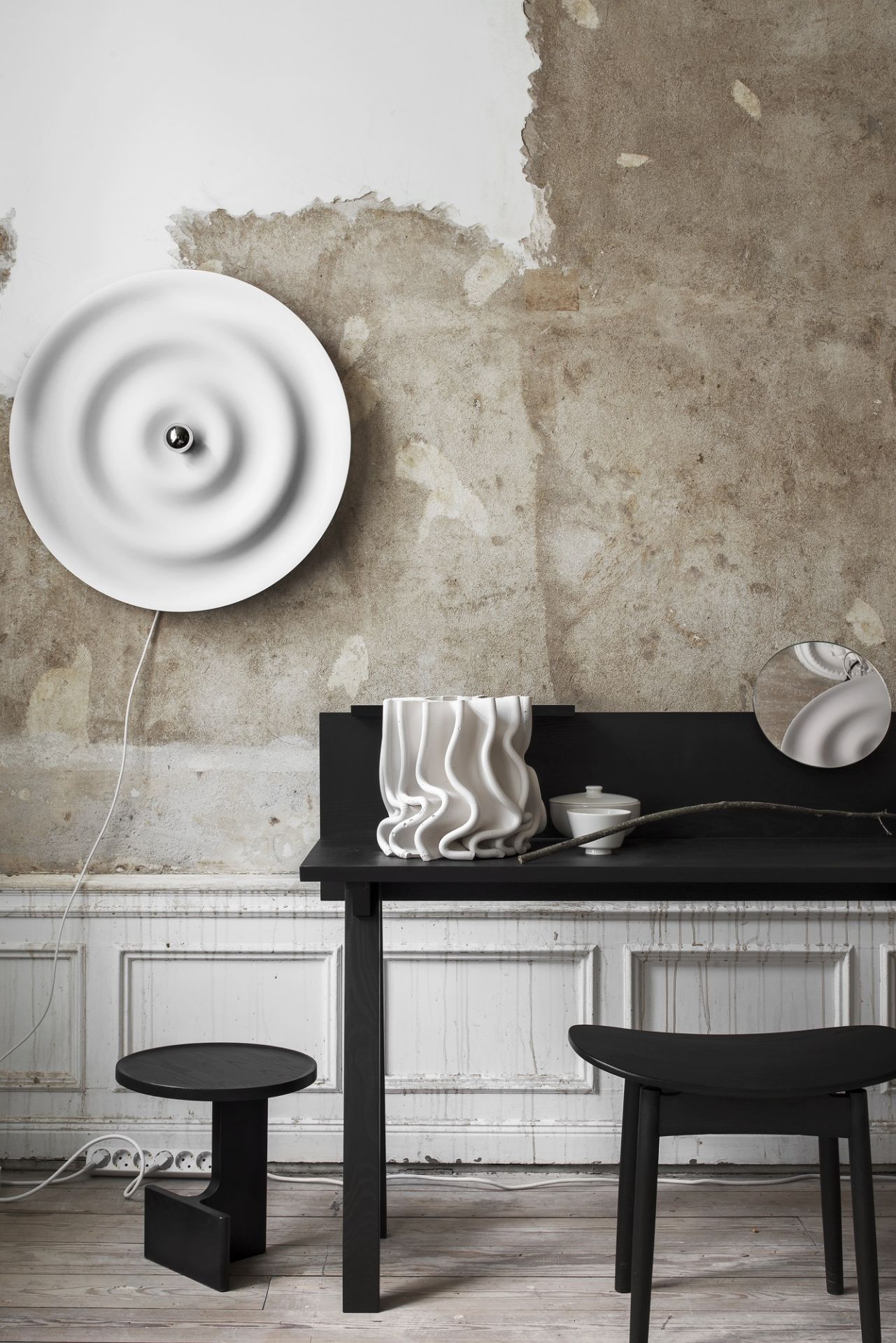 Weiße w171 Alma Wandleuchte von wästberg an einer Wand mit minimalistischer Tischdekoration und schwarzen Hockern.