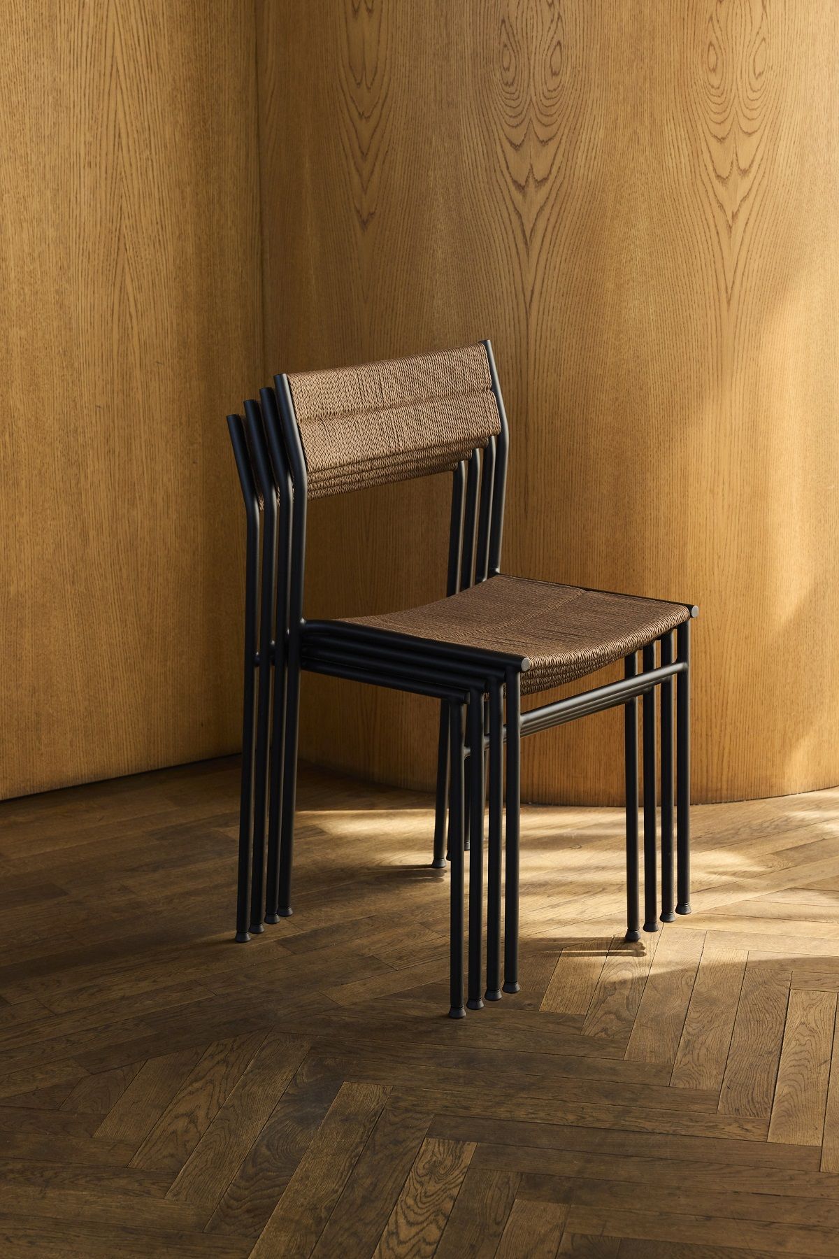 Gestapelte Vev Dining Stühle von Northern mit schwarzem Metallgestell und geflochtener Sitzfläche vor Holzwand.