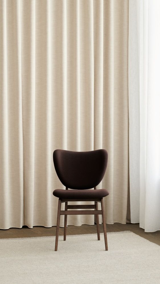Vollgepolsterter, brauner Elephant Dining Chair Stuhl von NORR11 vor beigen Vorhängen, geeignet für Esszimmer und Wohnbereich.