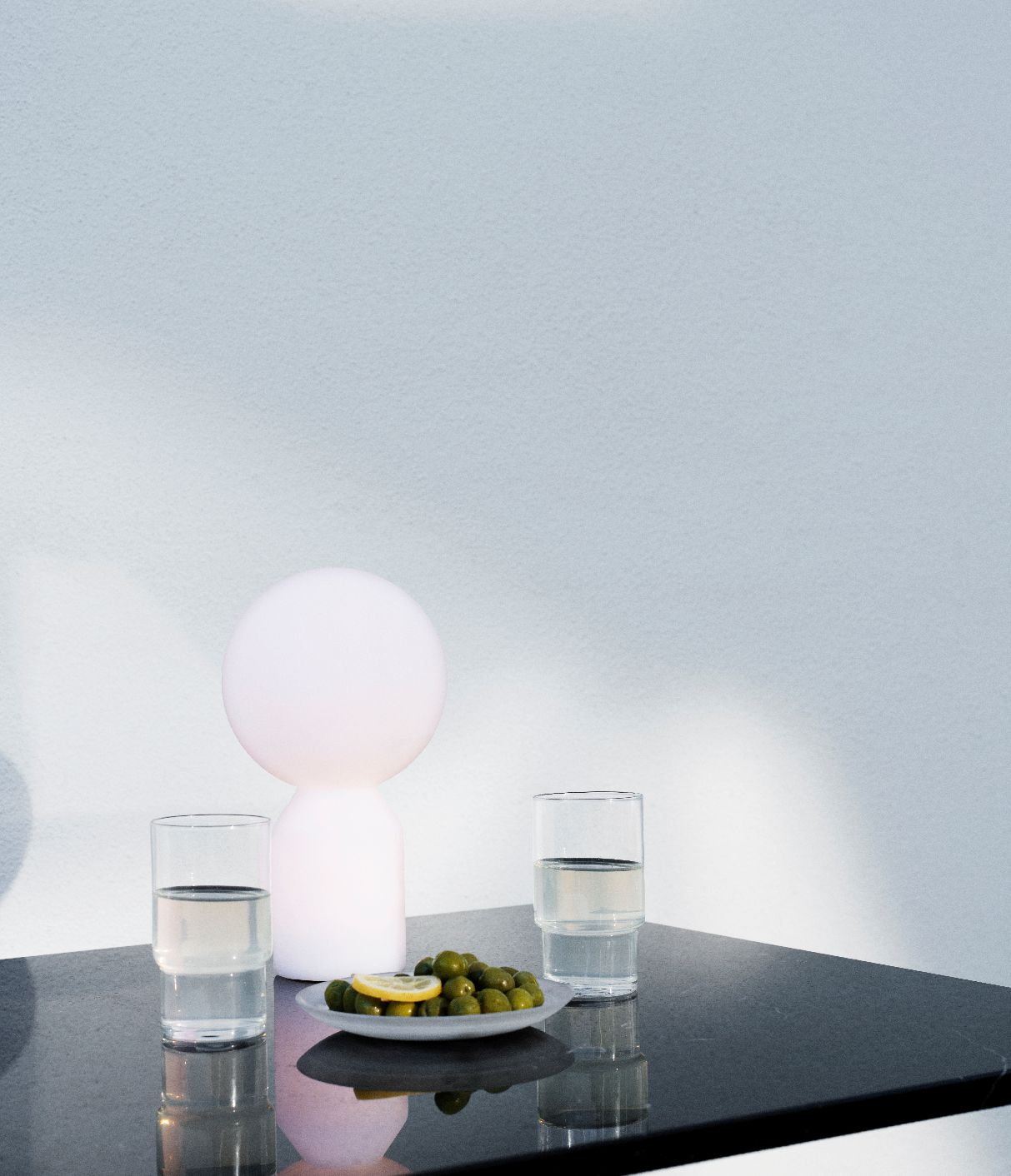 Nahaufnahme der Normann Copenhagen Yo Lamp Tischleuchte, weiß, neben Oliven und Wassergläsern auf dunkler Oberfläche.